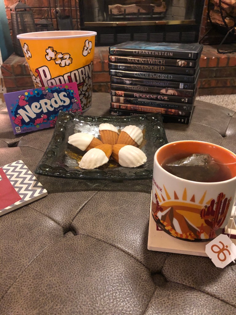 A stack of Halloween movies, with snacks including popcorn, candy, cookies, and a hot cup of tea.