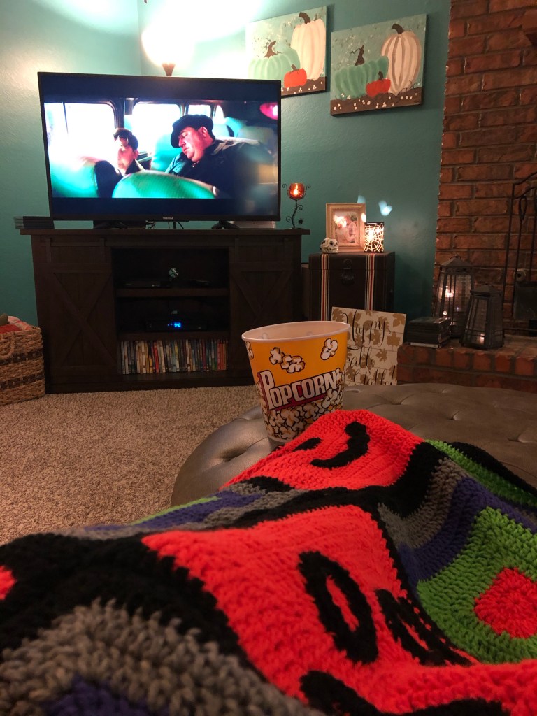 A view of the tv screen with a bucket of popcorn and a Halloween crocheted blanket in the foreground.